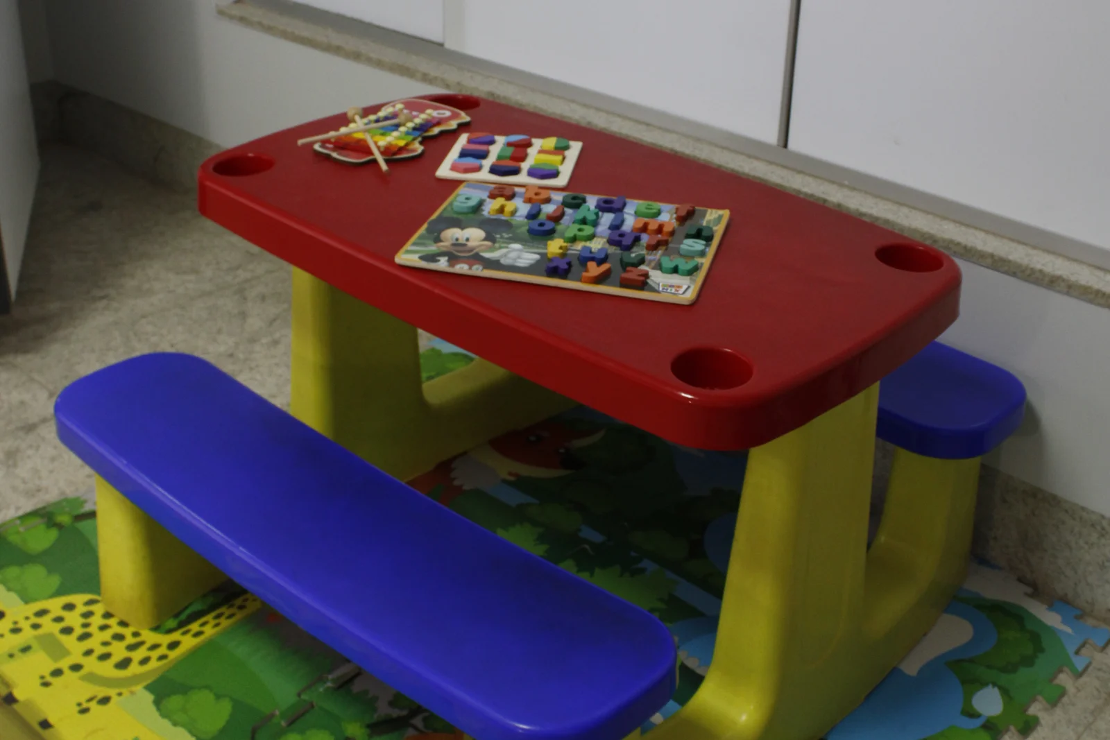 Mesinha e banco infantil sobre tapete de EVA colorido, no espaço com brinquedos da sala de aplicação